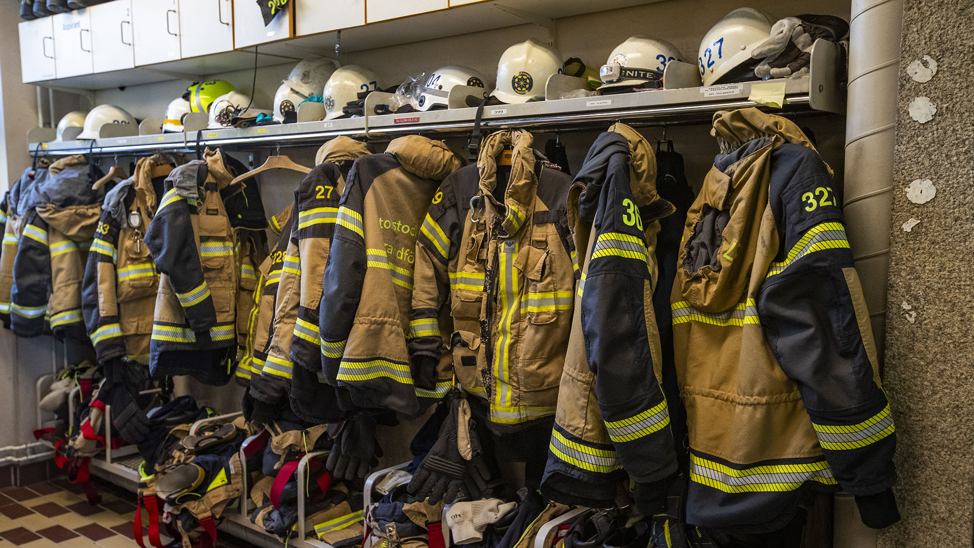 Brandmännens jackor, hjälmar, byxor och skor hänger längs väggen, foto.